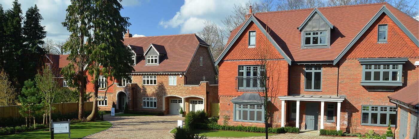 A view of a modern housing development featuring two large, detached red brick houses with tiled roofs. The house on the left has light-framed windows, a double garage, and decorative brickwork. The house on the right has grey-framed windows, a small balcony, and a covered entryway. Both houses have gabled sections and are set on landscaped lawns with paved driveways