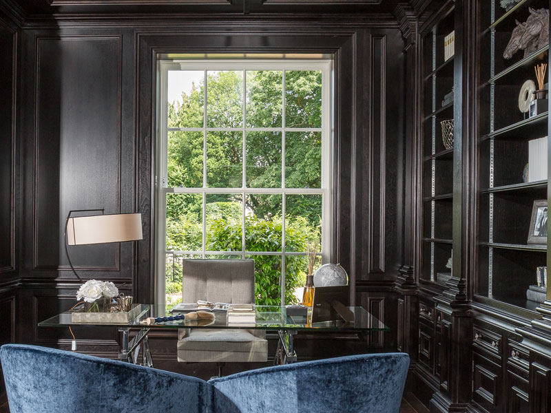 Luxurious office with a small library and glass desk and a white timber casement window behind the desk