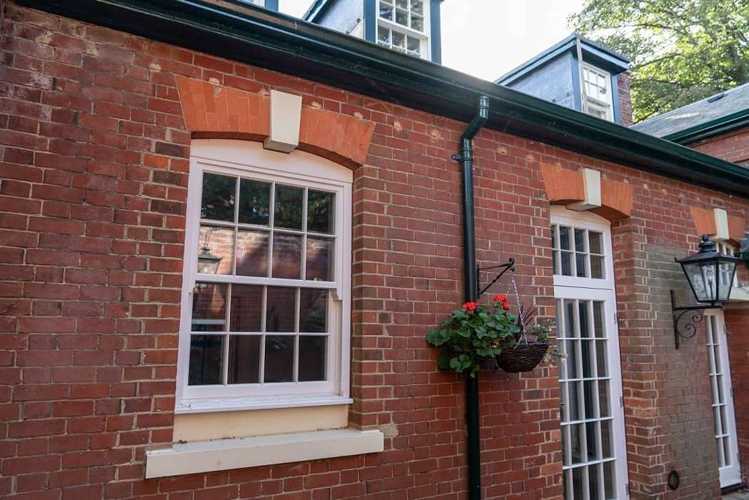 A detailed shot of the exterior of a red brick building with white timber sash windows. The window on the left has a pronounced white stone sill and a decorative arch of lighter-colored bricks and white stone details. A hanging basket with red flowers is suspended from a downpipe between the two windows. 