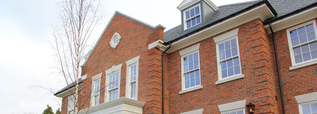 A three-story red brick building with multiple large, white timber windows. The windows are a mix of sash and casement styles, and some have decorative stone sills and lintels. The building has a pitched roof with dormer windows and a decorative gable. A single bare tree stands to the left of the building.