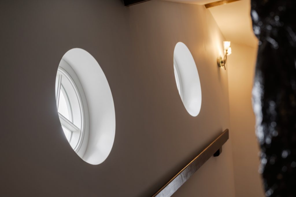 A low-angle shot captures two oval timber windows by Bereco set into a light-colored wall, illuminating a stairwell. A wooden handrail runs diagonally across the frame, leading up towards a warm wall light.