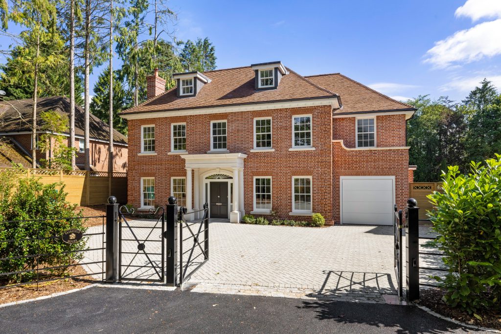 A large, traditional two-story red brick house with a tiled roof. The house has a symmetrical design, with a central white portico over a dark front door and several white timber sash windows on both floors. There are also dormer windows on the roof. The property is accessed through a black metal gate and features a paved driveway and an integrated garage. Tall trees are visible in the background.