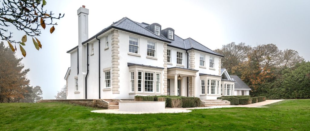 A large, white two-story house with a dark slate roof and numerous white timber sash windows. The house features decorative stone detailing around the windows and corners, a large central portico, and bay windows on either side. It is set on a grassy hill with trees in the background under a cloudy sky.