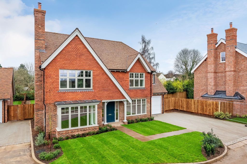 An elevated view of a large, newly built red-brick house with a prominent chimney and multiple Bereco timber casement timber windows. The house has a light-colored roof, a dark blue front door, and a well-maintained green lawn. 